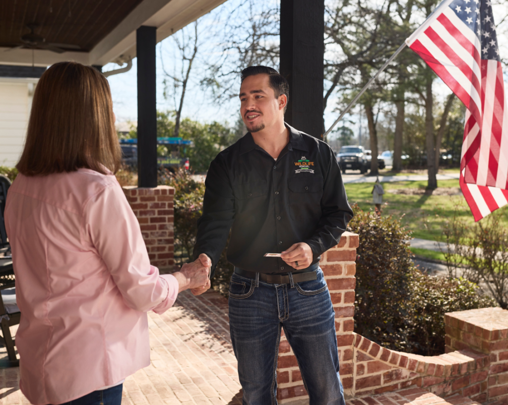 professional_expert Wildlife removal professional greeting a homeowner with a handshake on the front porch, standing beside an American flag and brick pillars, representing friendly and reliable service.